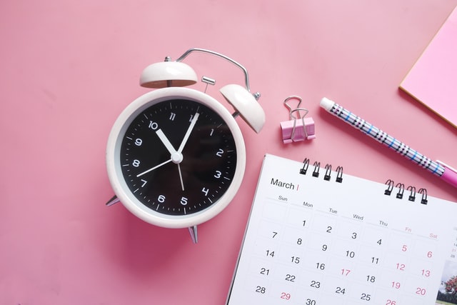 White alarm clock, pink paper clip, pen and white calendar with a baby pink background